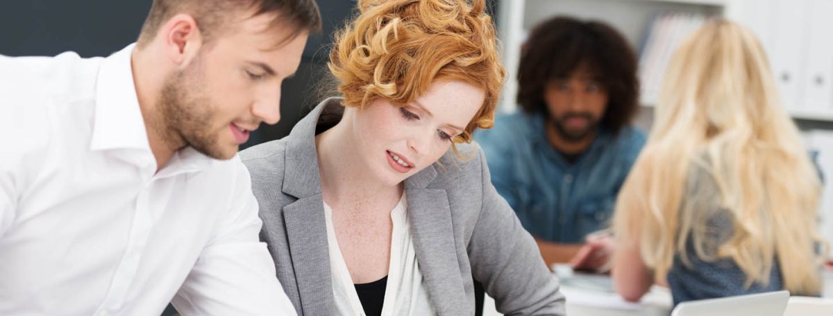 Business man and woman at work in the office pouring over paperwork on a desk together with multiethnic colleagues working in the background